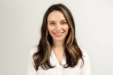 Sıcak ve cana yakın, uzun kahverengi saçlı ve hoş bir gülümsemesi olan Avrupalı genç bir kadın, beyaz bir bluz içinde yumuşak, tarafsız bir arka plan, stüdyo karşısında duruyor. İş, iş toplantısı.