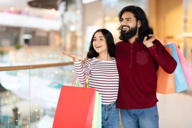 Heyecanlı genç Hintli çift alışveriş merkezinde duruyor, kucaklaşıyorlar ve gülümsüyorlar, boş fotokopi alanını gösteriyorlar, renkli kağıt poşetler alıyorlar, birlikte sezon alışverişinin keyfini çıkarıyorlar.