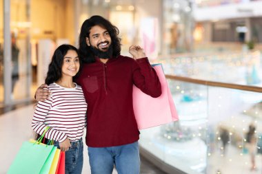 Güzel, genç Hintli çift alışveriş merkezinde duruyorlar, kucaklaşıyorlar ve gülümsüyorlar, boş fotokopi alanına bakıyorlar, renkli kağıt poşetler alıyorlar, birlikte sezon alışverişinin keyfini çıkarıyorlar.