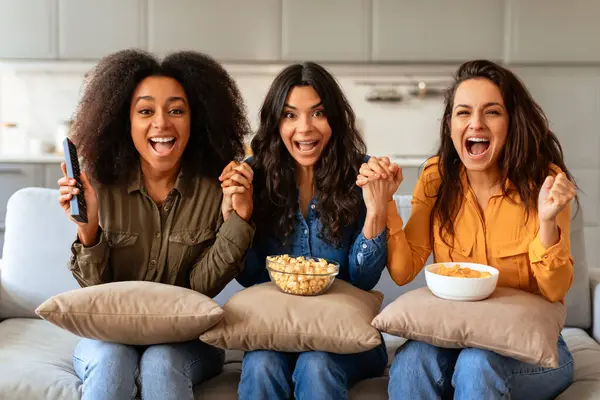 Three emotional diverse women cheerfully shouting reacting while watching TV indoor, cheering holding hands with excitement, sitting together on sofa at modern home interior with snacks
