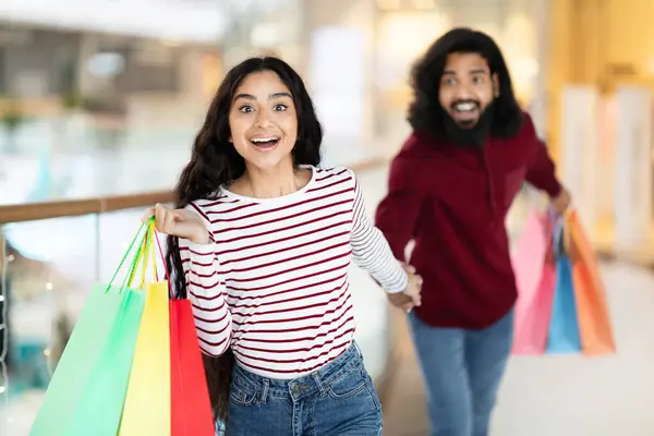 Excited attractive long-haired young indian woman wearing casual running by modern mall, pulling her boyfriend. Happy hindu couple carrying colorful paper bags purchases shopping together