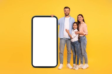 Geniş akıllı telefonlu aile, mesaj veya uygulama reklamı için boş ekran sunuyor. Ebeveynler ve kızları sarı arkaplanda telefon modeline yaslanıyorlar. Cep telefonu teklifini göstermek için mükemmel.