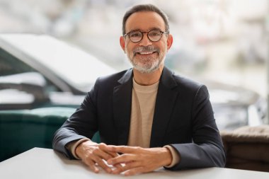 Kendinden emin ve arkadaş canlısı, gözlüklü, kameraya gülümseyen, modern bir kafe ortamında beyaz bir masada oturan, boş zamanların tadını çıkaran, video araması yapan Kafkasyalı kıdemli iş adamı.