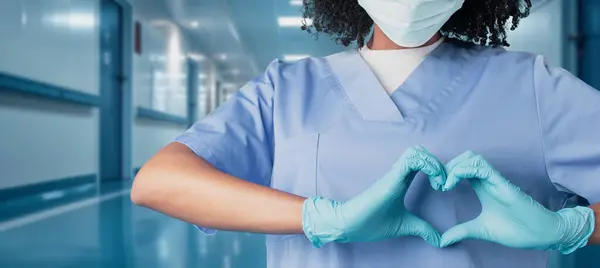一位戴着蓝色刷子顶盖和防护面罩的医护人员 用戴着手套的手塑造了心脏 传达了在诊所 全景医疗领域的爱与关怀信息 — 图库照片