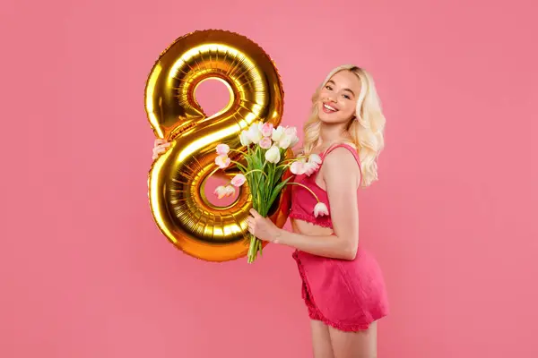 金8バルーンとピンクのチューリップを保持する魅力的なブロンドは 遊び心のある服を着て ピンクの背景に喜びとWomens Dayのお祝いを体現しています — ストック写真