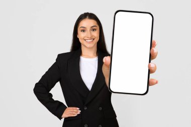 Siyah ceketli, kendine güvenen genç iş kadını boş bir akıllı telefon ekranı sunuyor. Tarafsız arka planda sıcak, davetkar bir gülümseme, iş uygulamasının bir kopyası.