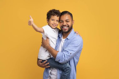 Babası onu tutarken gülümseyen siyah çocuk, mutlu Afrikalı genç babası ve küçük oğlu sarı stüdyo arka planında izole edilmiş poz vererek birlikte eğleniyorlar.