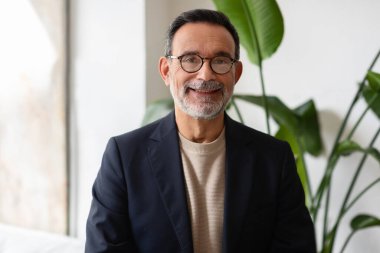 Gözlüklü, sıcacık gülüşlü, spor ceket ve balıkçı yaka kazak giymiş, arka planda tropikal bitkilerle dolu bir ofiste duran, kendinden emin, beyaz, kıdemli bir işadamı.