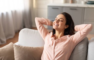 Memnun ve huzurlu bir kadın, elleri başının arkasında uzanmış, kanepeye uzanmış, mutlu bir ifadeyle, parlak bir odada.