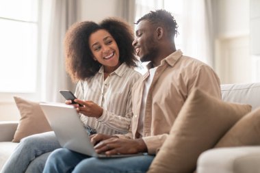 Dijital bağlantı. Mutlu siyah milenyum kocası dizüstü bilgisayar kullanırken, karısı akıllı telefonuna göz gezdirirken, çiftler aletlerle internet keyfi yaparken, evde oturma odasında, modern iletişime dalmışken,