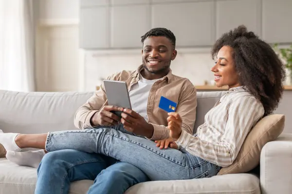 Tech savvy buyers. Happy African american couple holding credit card exploring webstore on tablet computer, making digital purchase and paying online while shopping in modern living room