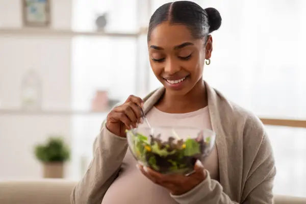 Diet and nutrition during pregnancy. Portrait of happy young african american pregnant woman wearing comfy casual clothing sitting on couch, eating healthy vegetable salad at home, copy space