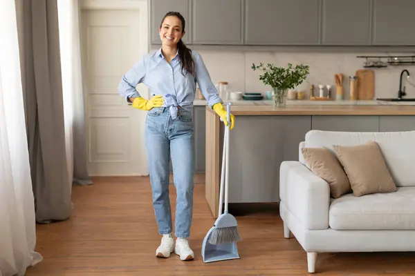 Happy woman in casual attire and yellow cleaning gloves standing with broom, posing to start her household chores in modern kitchen, full length, free space