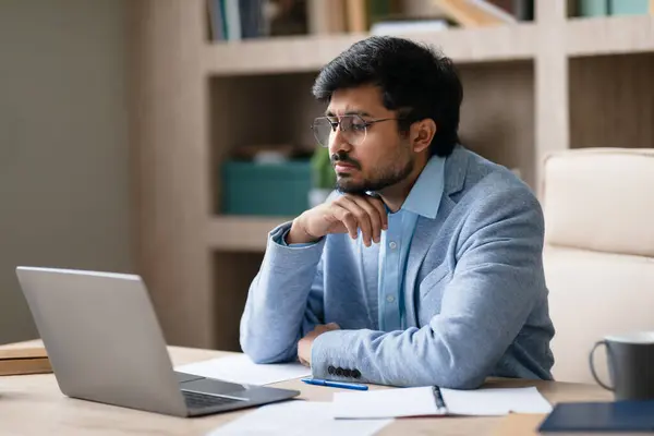 Thoughtful Indian professional businessman pondering strategies while reviewing work on his laptop in contemporary office environment, thinking over problem solutions at workplace