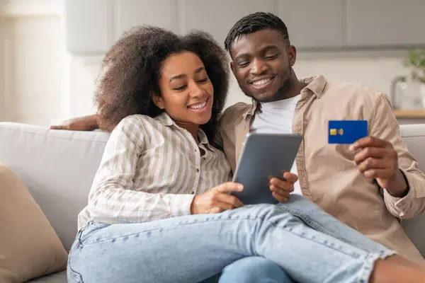 Online shopping joy. Happy black millennial couple enjoying digital retail on tablet, holding credit card for easy internet payments, browsing webstores on sofa at home. Modern ecommerce