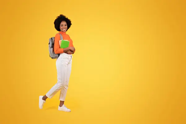 年轻的非洲裔美国女人 留着自然的头发 穿着橙色的高领毛衣和白色裤子 自信地摆出一副灰色背包和绿色笔记本的架势 衬托着充满活力的黄色背景 — 图库照片