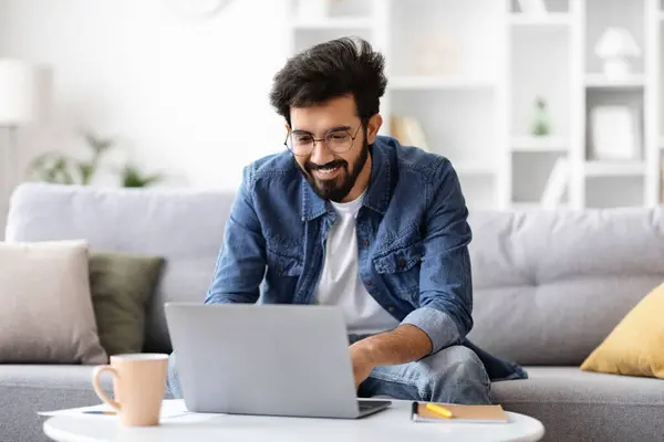 Cheerful indian man working on laptop and enjoying cup of coffee at home, happy young eastern male freelancer using computer, comfortably sitting at desk in bright modern living room, free space