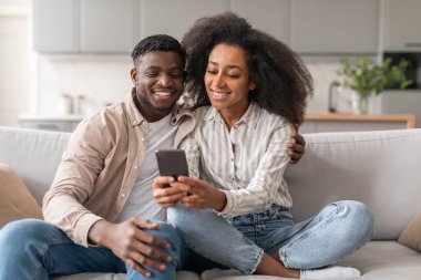 Kanepede oturan neşeli Afro-Amerikan çift evde akıllı telefonlarıyla eğlenirken, sosyal medya haberleri sunuyor, sohbet ediyor ve video izliyorlar. Modern eğlence ve dijital yaşam tarzı