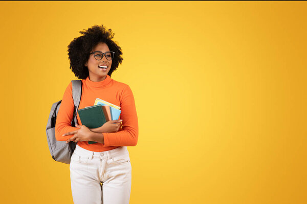 Cheerful African American young lady with glasses and curly hair, holding colorful books, wearing an orange sweater, white pants, and a grey backpack, looking away on a yellow background