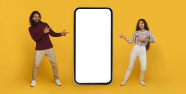 Güzel online teklif. Havalı genç Hintli erkek ve kadın büyük akıllı telefonu işaret ediyor aralarında beyaz ekran maketi kopyalama alanı var, sarı arka plan, web-pankart