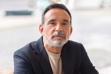 Düşünceli bir ifadeyle, siyah ceket ve bej bir balıkçı yaka kazak giyen, parlak, havadar bir odada, tuzlu ve biberli sakallı, hazır ve nazır bir beyaz adamın yakın çekimi.