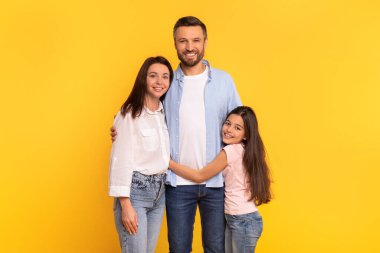 Üç kişilik neşeli bir aile, anne, baba ve çocuk sarı arka planda kucaklaşıyorlar, birlikteliğin ve aile bağının tadını çıkarıyorlar, kameraya gülümseyen sevgi ve mutluluğu iletiyorlar.