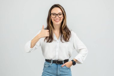 Gözlüklü, uzun saçlı, gülümseyen Avrupalı bir kadın kameraya gülümserken beyaz bluz, kot pantolon ve akıllı bir şekilde başparmağını kaldırıyor.