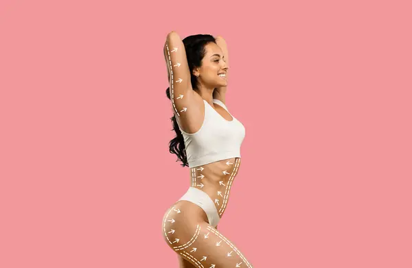 Smiling young brunette woman in white underwear with arms behind her head and white massage lines on her slim body posing on pink background, copy space. Body shaping, sculpting treatment