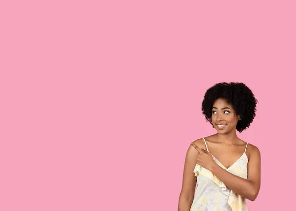 一个快乐的千年非洲裔美国女人 卷曲的头发 身穿淡淡的花裙 站在旁边 展示着一种产品或想法 粉色背景上有着滑稽的表情 — 图库照片