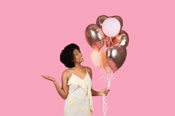 一个快乐的千年非洲裔美国女人 卷曲的头发 手里拿着金色的玫瑰和白色的气球 面带微笑地站在粉色的背景下 — 图库照片