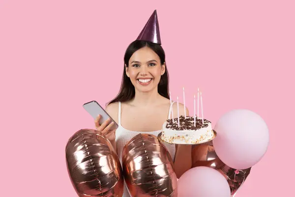 パーティー帽子の放射状の若い女性は ライトキャンドルと光沢のある風船で誕生日ケーキを開催し ピンクの背景に彼女のスマートフォンで陽気なポーズ — ストック写真