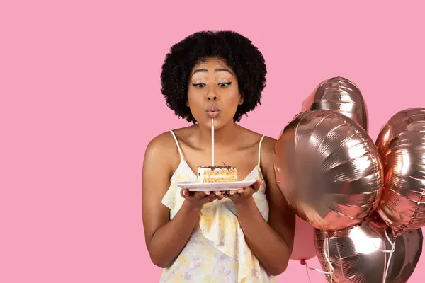Delighted millennial African American woman with curly hair blowing out a candle on a slice of birthday cake, with rose gold balloons in the background, on a pink backdrop