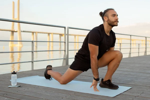 Outdoor fitness. Motivated young man in sporty attire doing leg stretches on mat outside. Guy making kneeling hip flexor exercise during stretching training on pier. Workout, wellbeing