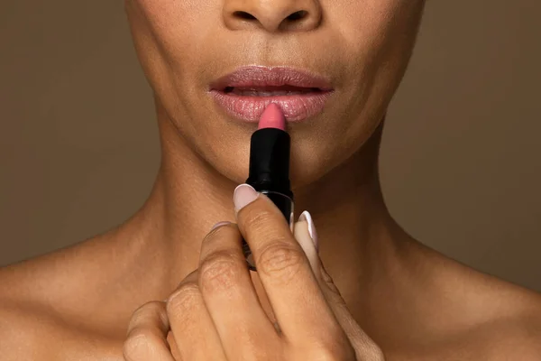 Closeup of black middle aged woman applying bright pink lipstick, focusing on her lips and the tip of the lipstick held by her manicured fingers, cropped