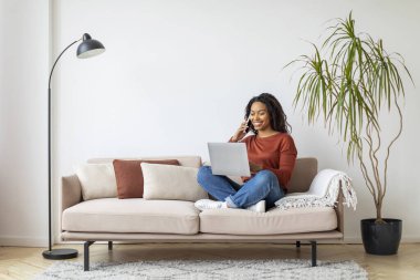 Çevrimiçi Çalışma Konsepti. Genç siyahi kadın dizüstü bilgisayarla çalışıyor ve cep telefonuyla konuşuyor, Afrikalı Amerikalı kadın bilgisayarını serbest çalışmak için kullanıyor, oturma odasında koltukta oturuyor, uzayı kopyalıyor.