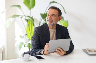 Olgun Kafkas iş adamının elinde tablet ve neşeli bir ifadeyle masada bir fincan kahve ve akıllı telefonla ofis yeşiliyle çevrili olması iyimser bir sevinç.
