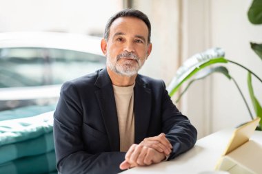 Kendine güvenen, düzgün kesilmiş sakallı, aydınlık bir ofiste rahatça oturan, elleri açık bir defter ve beyaz seramik bir fincanın yanındaki masaya yapışan üst düzey bir yönetici.