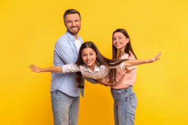 Neşeli beyaz anne-babalar küçük kızlarını kaldırırken, kız kollarını uçak kanatları gibi açıyor, sarı stüdyo arkaplanında eğleniyor. Seyahat ve tatilin neşesi