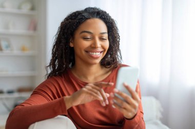 Evde cep telefonu kullanan mutlu siyahi kadının portresi, akıllı telefondan alışveriş yapan veya arkadaşlarıyla mesajlaşan gülümseyen Afro-Amerikan bayan, koltukta dinlenirken yeni uygulamalara göz gezdirirken...