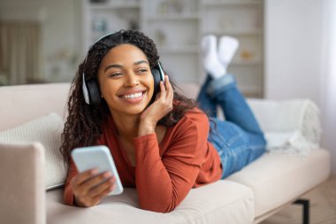 Yurtiçi rahatlama. Gülümseyen Siyahi Kadın Portresi, Akıllı Telefonla Müzik Dinleyen Mutlu Afro-Amerikan Kadın Kulaklık Takan Kanepede uzanan Favori Şarkılar, Kapanış