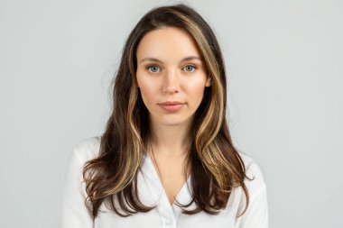 Uzun esmer saçlı, beyaz bluzlu, sakin ve yaklaşılabilir bir ifadeyle doğrudan kameraya bakan sakin, genç Avrupalı bir kadının portresi.