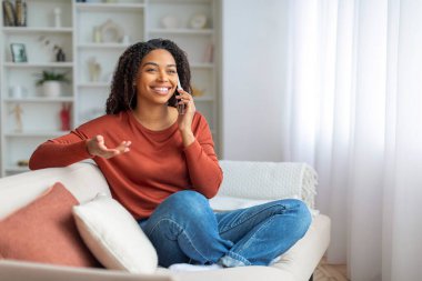Evde cep telefonuyla konuşan mutlu Afrikalı Amerikalı kadın portresi, oturma odasında koltukta otururken keyifli cep telefonu sohbetleri yapan neşeli siyah bayan,