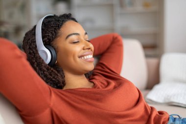 Güzel, kablosuz kulaklıklı, gülümseyen siyah kadın evde müzik dinliyor, genç, Afro-Amerikan bayan arkana yaslanıp kapalı gözlerle koltuğa yaslanıyor, favori müzik listesinin tadını çıkartıyor,