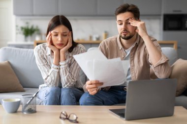 Endişeli genç Avrupalı çift finansal belgeleri dizüstü bilgisayarla inceliyor. Oturma odasındaki kanepede stres ve tefekkür belirtileri gösteriyor.