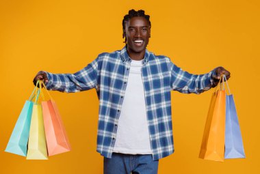 İki elinde renkli alışveriş torbaları tutan neşeli, genç siyahi adam. Ekose gömlek giyen Afro-Amerikan adam. Sarı stüdyo arka planında tek başına duruyor.