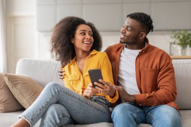 Afrika kökenli Amerikalı çiftler akıllı telefondan başvurularını kullanıyorlar, eşleri cep telefonlarını kocalarına gösteriyorlar birlikte eğleniyorlar, evdeki kanepede oturuyorlar. İletişim, bağlı yaşam tarzı