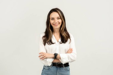 Dostça gülümseyen, kolları çapraz duran, beyaz bluz, mavi kot pantolon ve arka planda akıllı bir kadın.