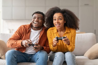 Neşeli siyah milenyum çifti evlerinde oyun konsolunda oyun oynuyorlar, ev eğlencesi ve dijital eğlencenin tadını çıkarıyorlar, ortak hobi aktivitelerinin eğlencesini sergiliyorlar.
