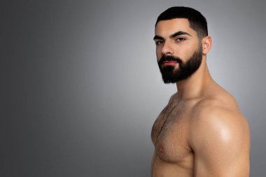 Erkekler için bahşişler önemlidir. Yakışıklı, atletik, çıplak esmer, orta doğulu, kalın sakallı genç adam kameraya bakıyor, gri stüdyo arka planında izole edilmiş, kopyalama alanı var.