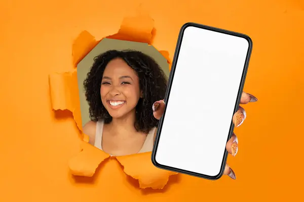 快乐的非洲裔美国女人带着喜形于色的笑容 拿着装有空白屏幕的智能手机 在撕破的橙纸中窥视着 适于设计模型或广告 — 图库照片
